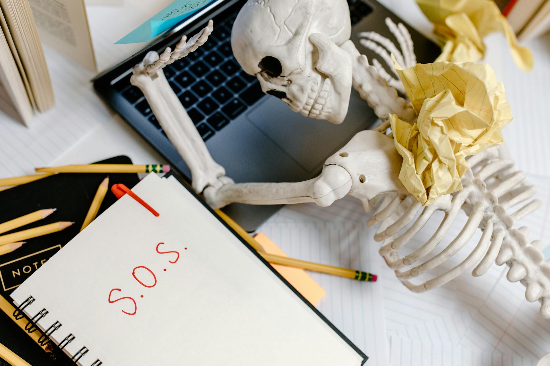 Skeleton slumped over cluttered desk with crumpled paper and notebook reading S.O.S., symbolizing burnout, work overload, and creative exhaustion at laptop
