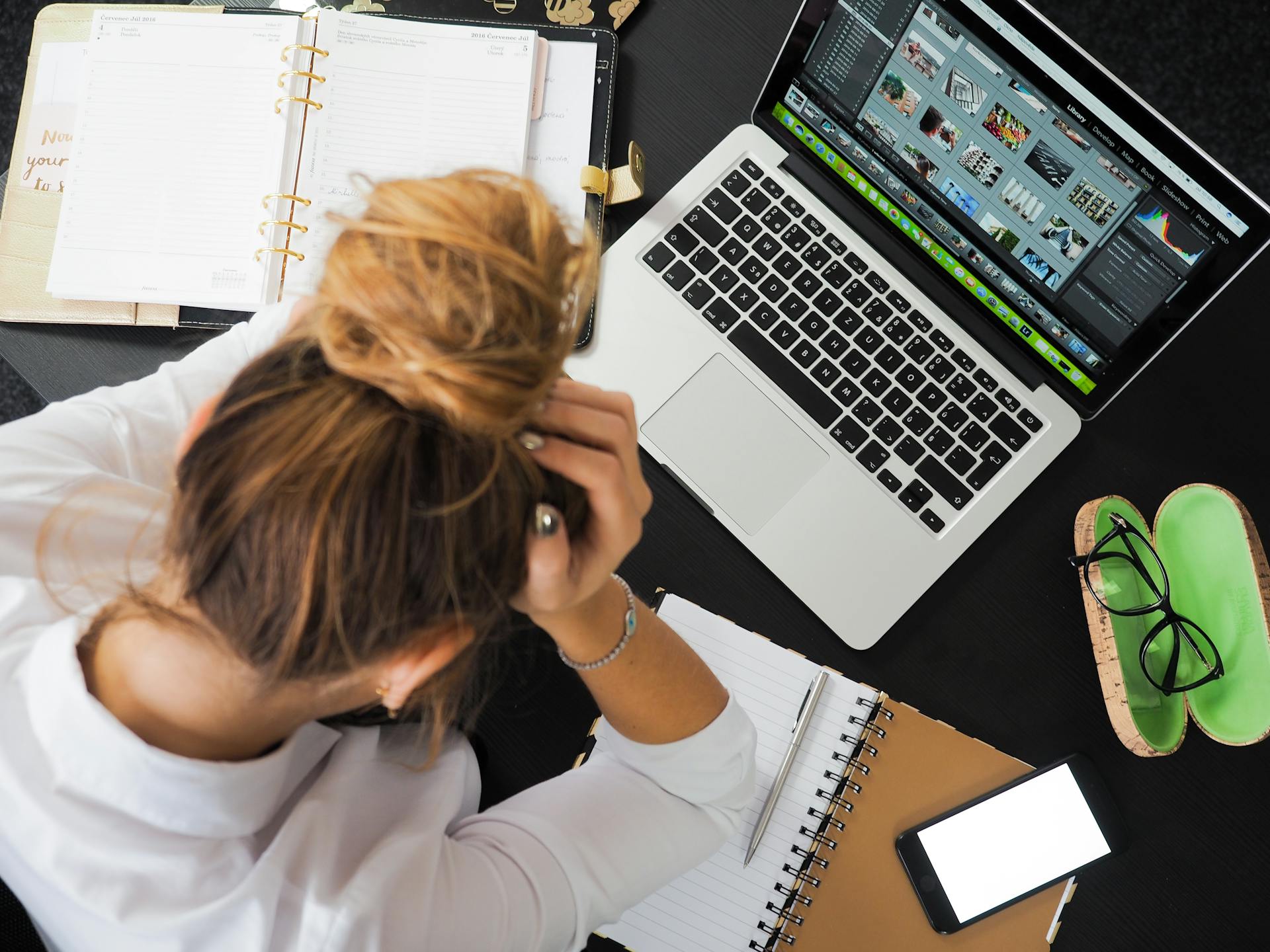 Overwhelmed woman clutching head at cluttered desk with laptop, planner, and phone—illustrating digital burnout, design fatigue, and ecommerce workflow stress